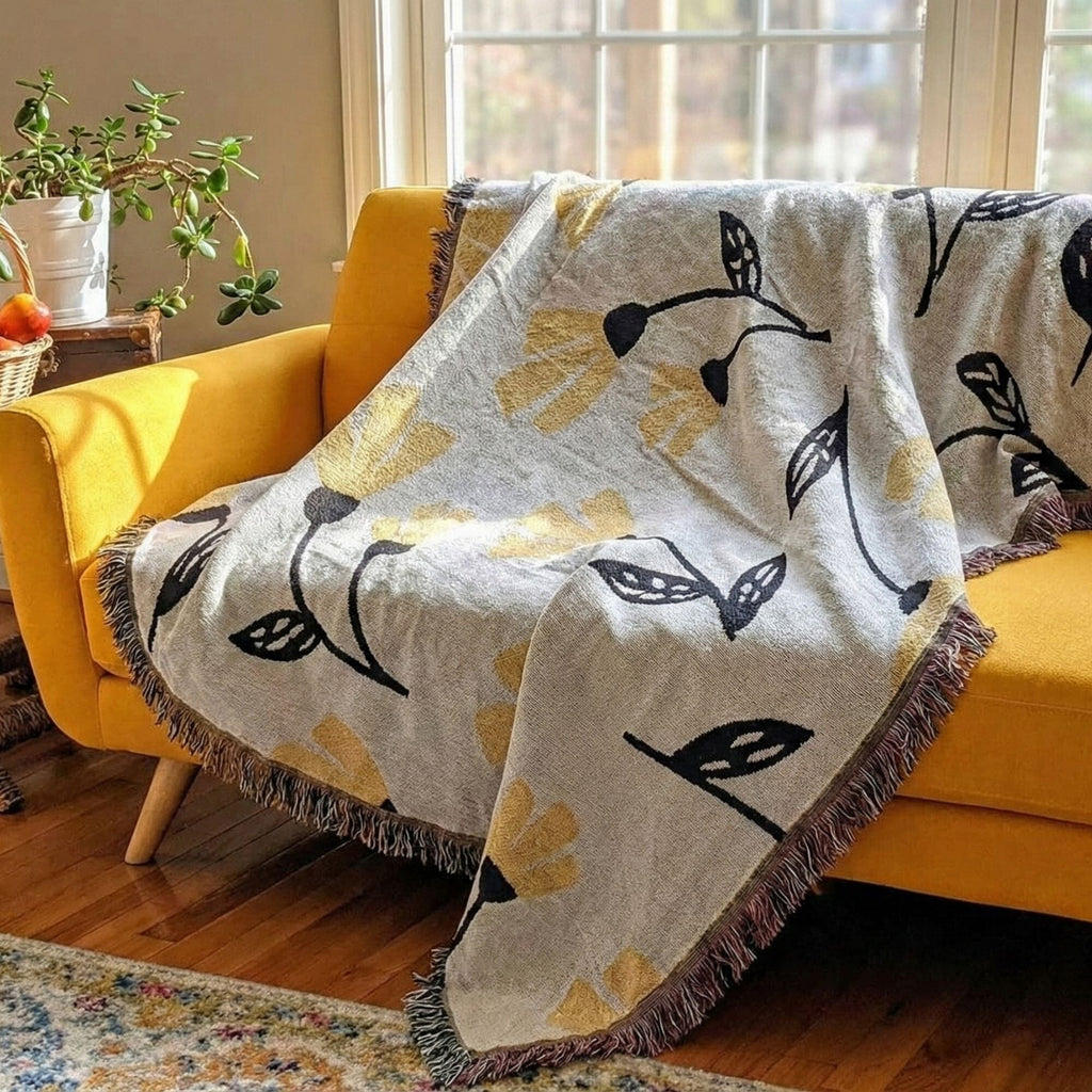 A woven throw blanket with a black and yellow floral vine pattern and fringed edges draped over a mustard yellow armchair in a bright living room.